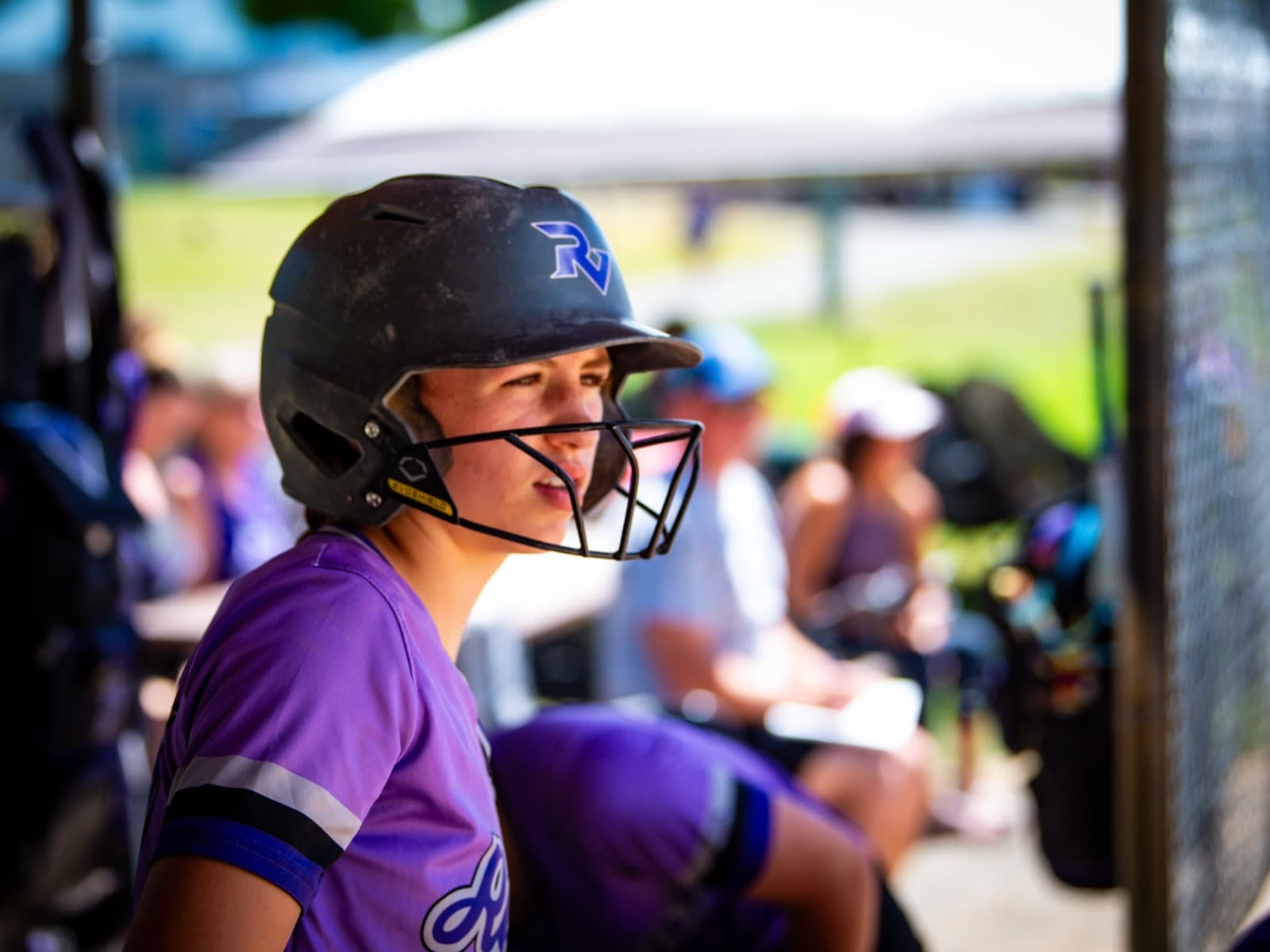 A Ravens softball player at the dugout rail, helmet on, watching the game